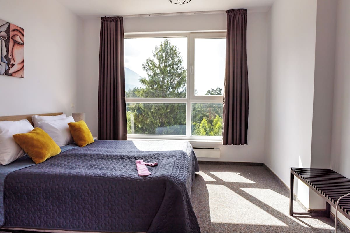 A bedroom with a bed, a window with curtains, a bench, and a painting on the wall.