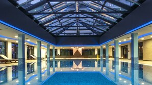 An indoor swimming pool with a glass ceiling, surrounded by lounge areas and a staircase leading to upper levels.
