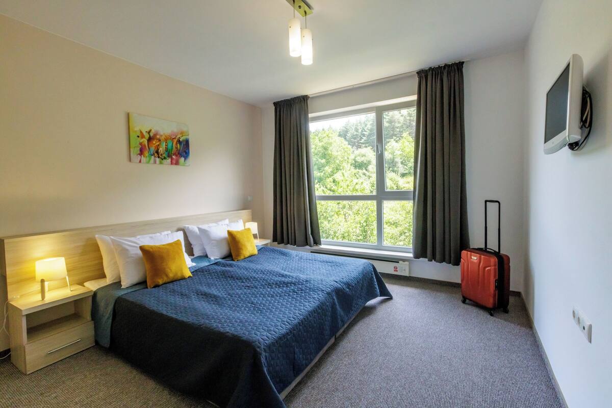 A hotel room with a bed, a nightstand, a lamp, a suitcase, a window with curtains, and a painting on the wall.
