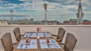 A terrace with wicker chairs and tables, a vase with flowers, and a view of a cityscape and water.