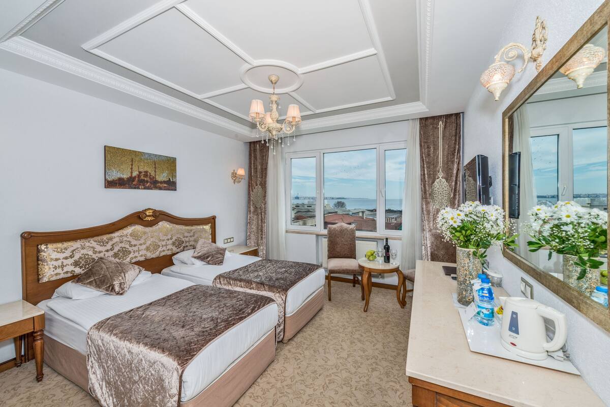 A hotel room with two beds, a sitting area with a view, a large mirror, and a chandelier.