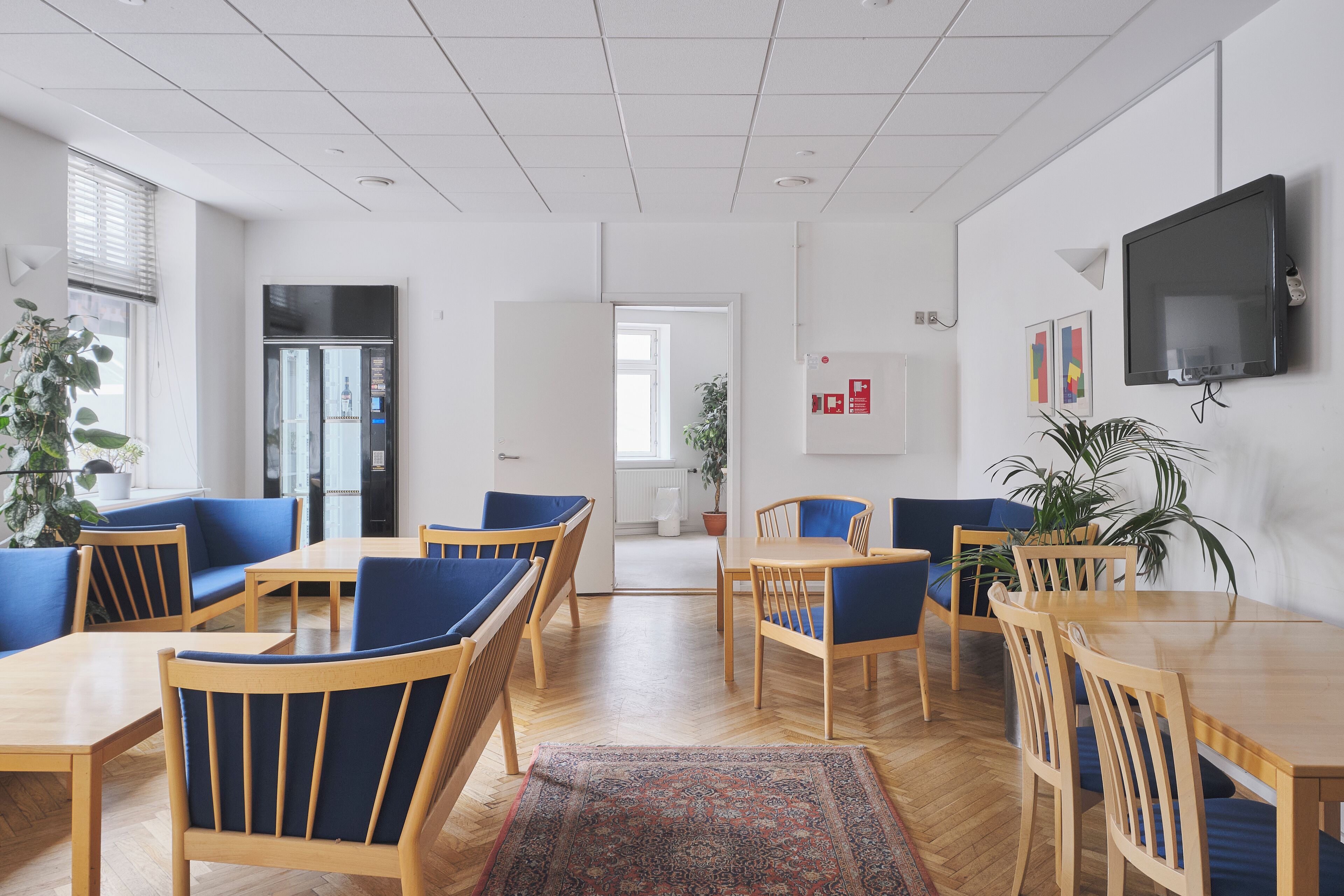 Ein zeitgemäßer Essbereich mit Holztischen und blau gepolsterten Stühlen, ein großer Flachbildfernseher an der Wand und eine Zimmerpflanze auf der rechten Seite.