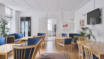 Ein zeitgemäßer Essbereich mit Holztischen und blau gepolsterten Stühlen, ein großer Flachbildfernseher an der Wand und eine Zimmerpflanze auf der rechten Seite.