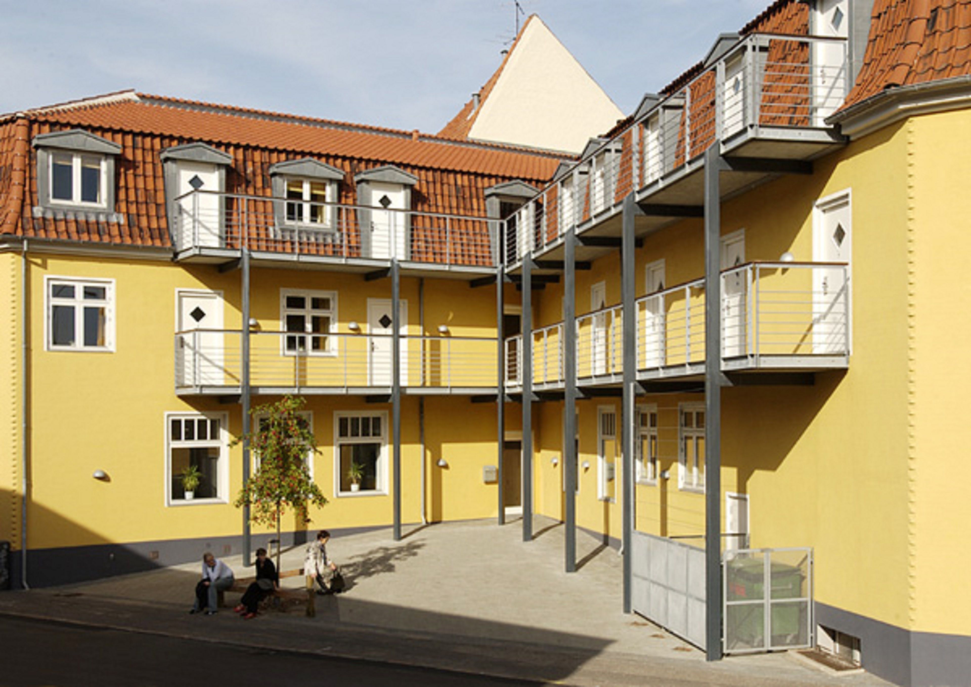 A yellow multi-story building with balconies, a central courtyard, and a small tree.