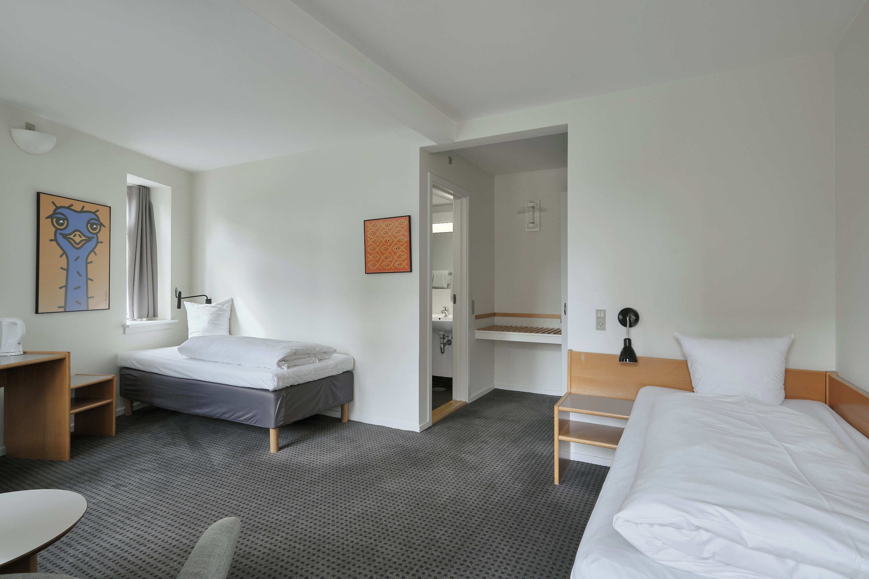 A hotel room with a bed, a desk, a chair, a small table, and a bathroom visible through an open door.