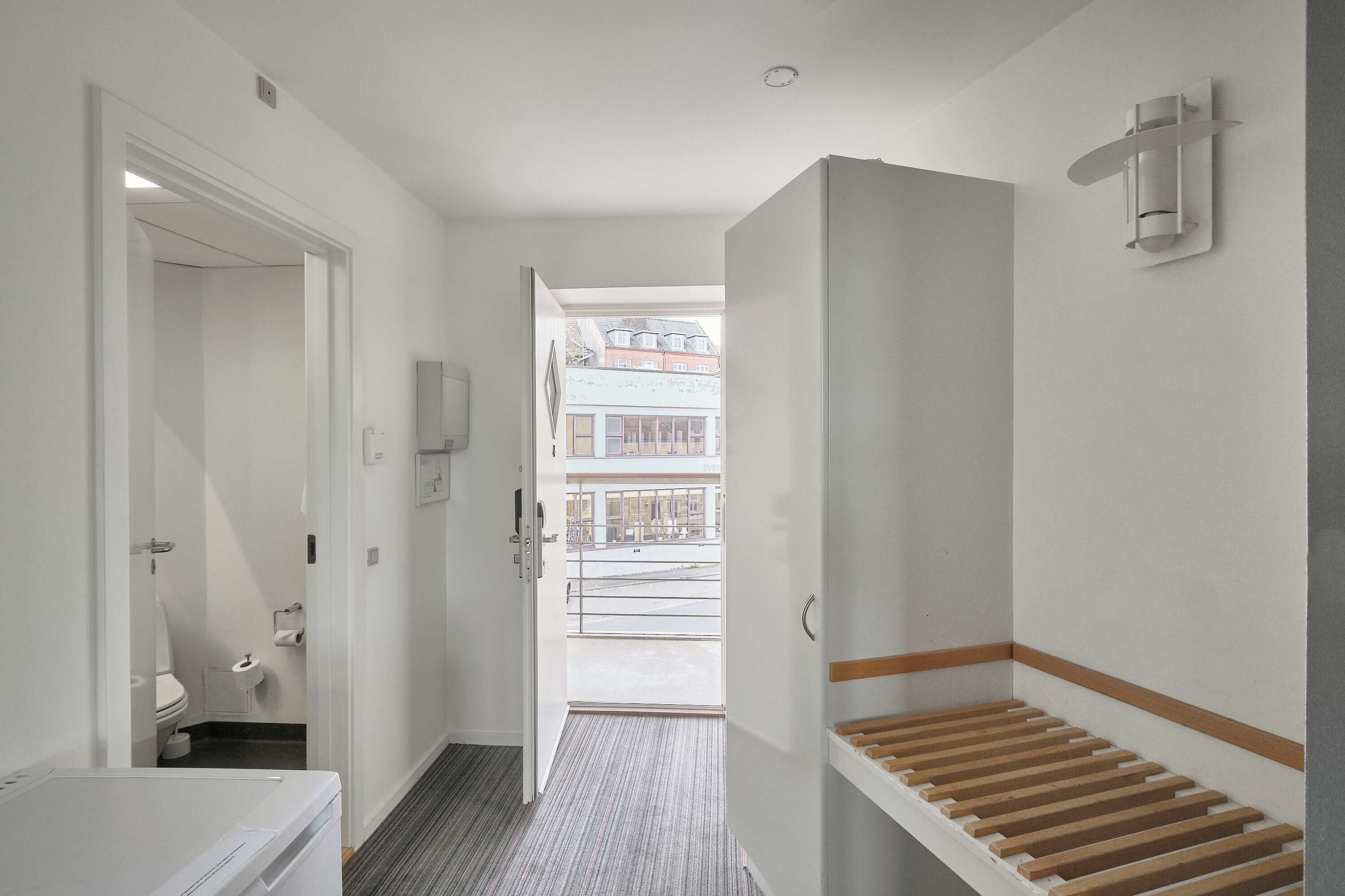 A hotel room with a bathroom, a wooden bench, and a view of buildings outside.