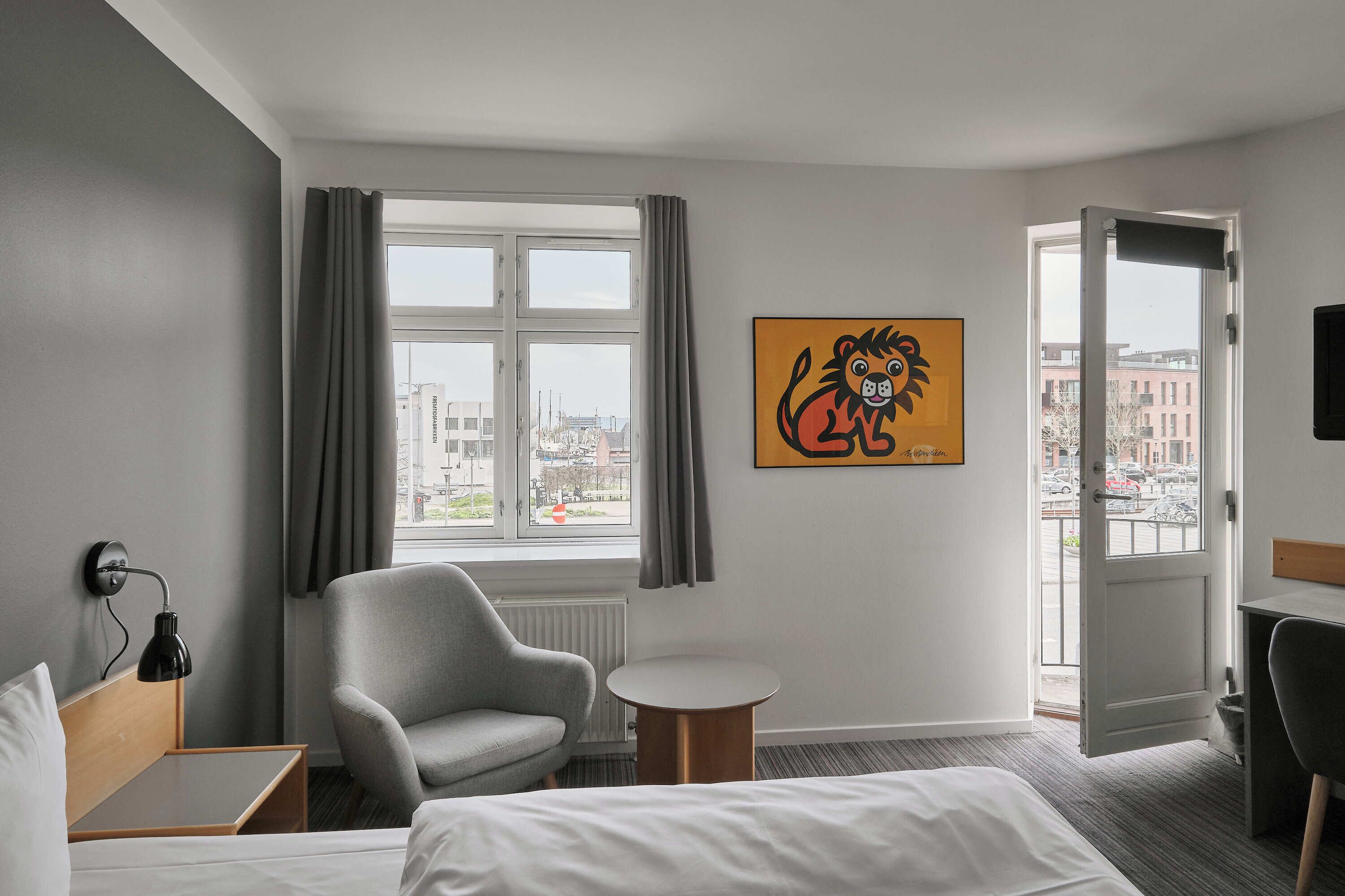 A hotel room with a bed, a grey armchair, a small round table, a window with curtains, and a city view.