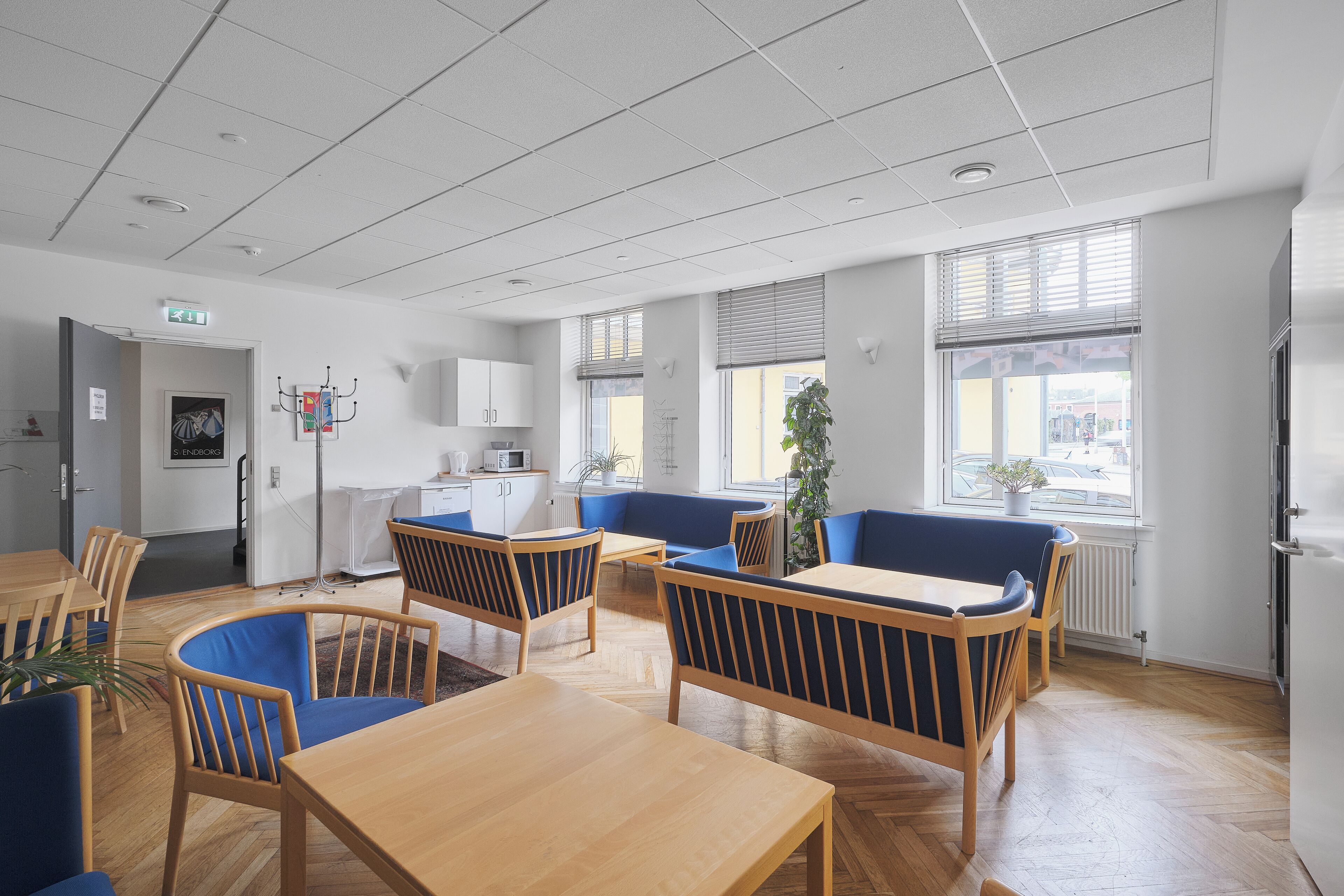 A modern dining area with wooden tables and blue cushioned chairs, a kitchenette in the background, and large windows providing natural light.