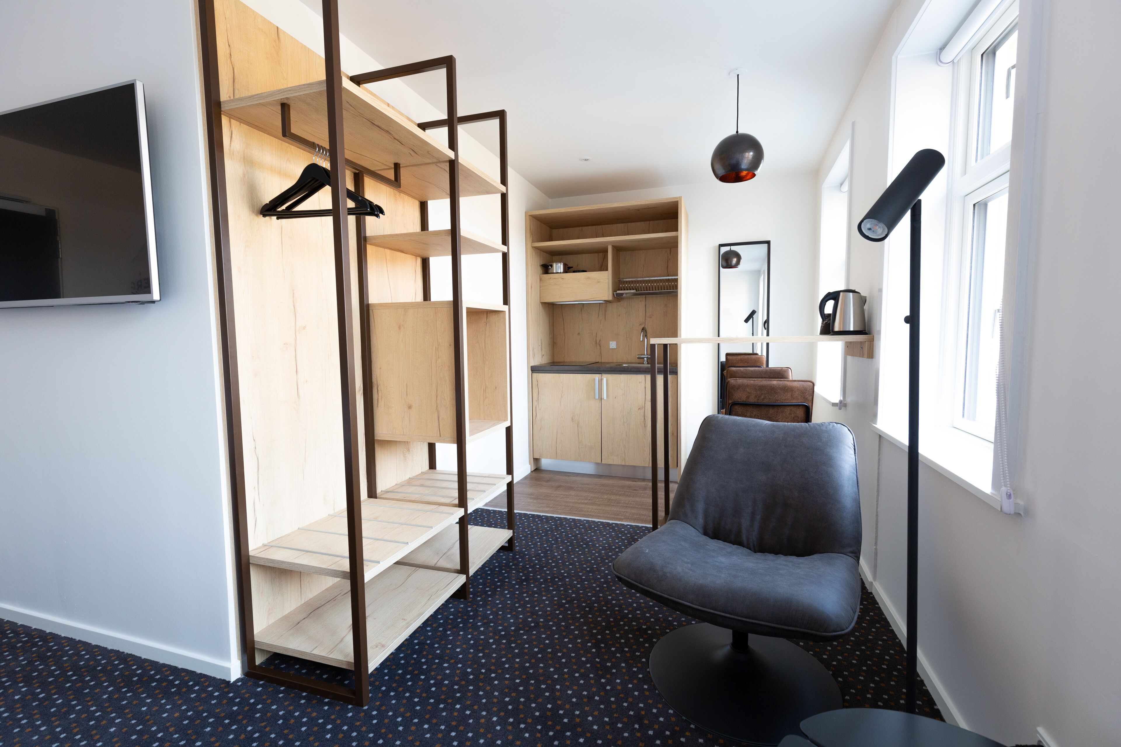 A modern, compact room with a built-in wardrobe, a seating area, a small kitchen, and a large window providing natural light.