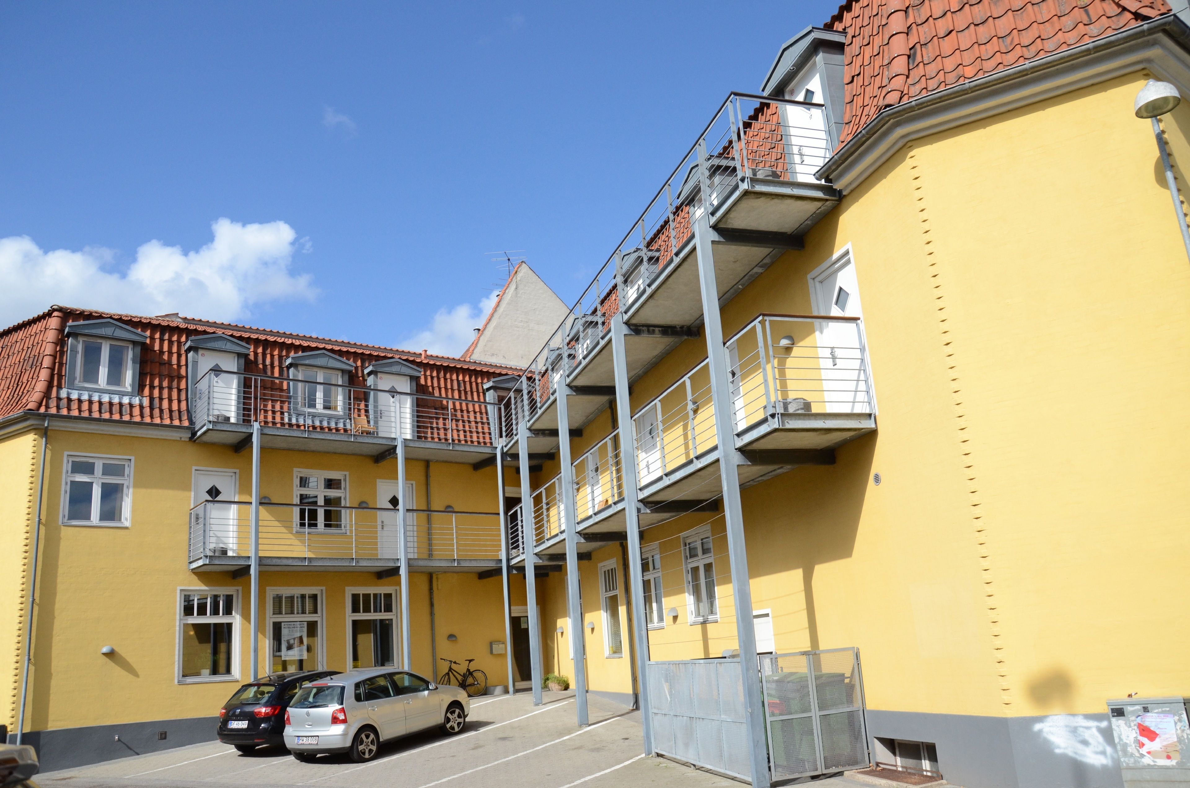 A yellow building with a red-tiled roof, multiple balconies, and a parking area with cars.