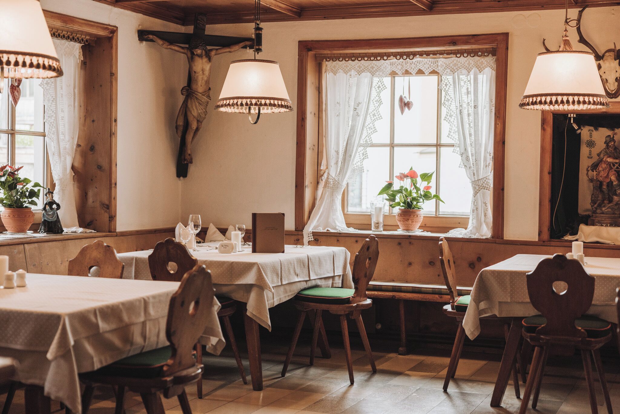 A dining area with wooden tables and chairs, a crucifix on the wall, and a window with lace curtains.