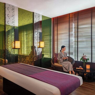 A modern hotel room with a large bed, a seating area with a table, and a view through large windows with blinds.