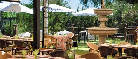 Un área de comedor al aire libre con mesas dispuestas para dos, rodeada de sillas de mimbre y cubierta por sombrillas blancas. Una fuente decorativa se encuentra en el centro.