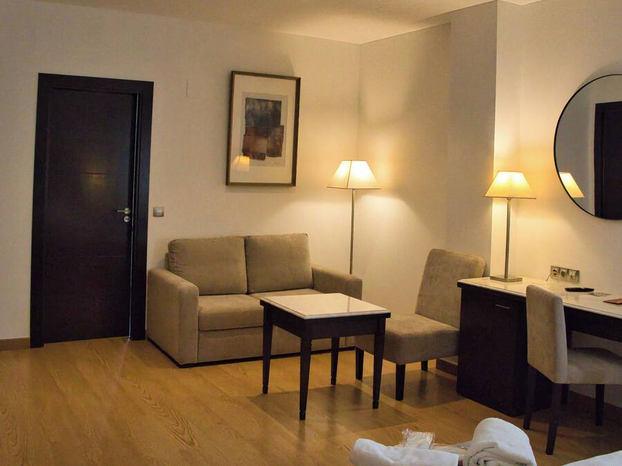 A hotel room with a sofa, a small table, a desk with a chair, a lamp, and a mirror.
