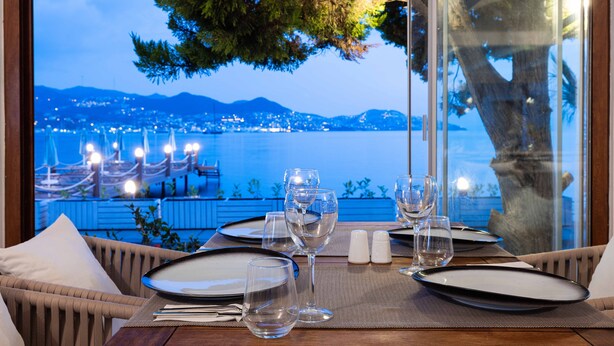 A dining table set for two with a view of a coastal city at dusk.