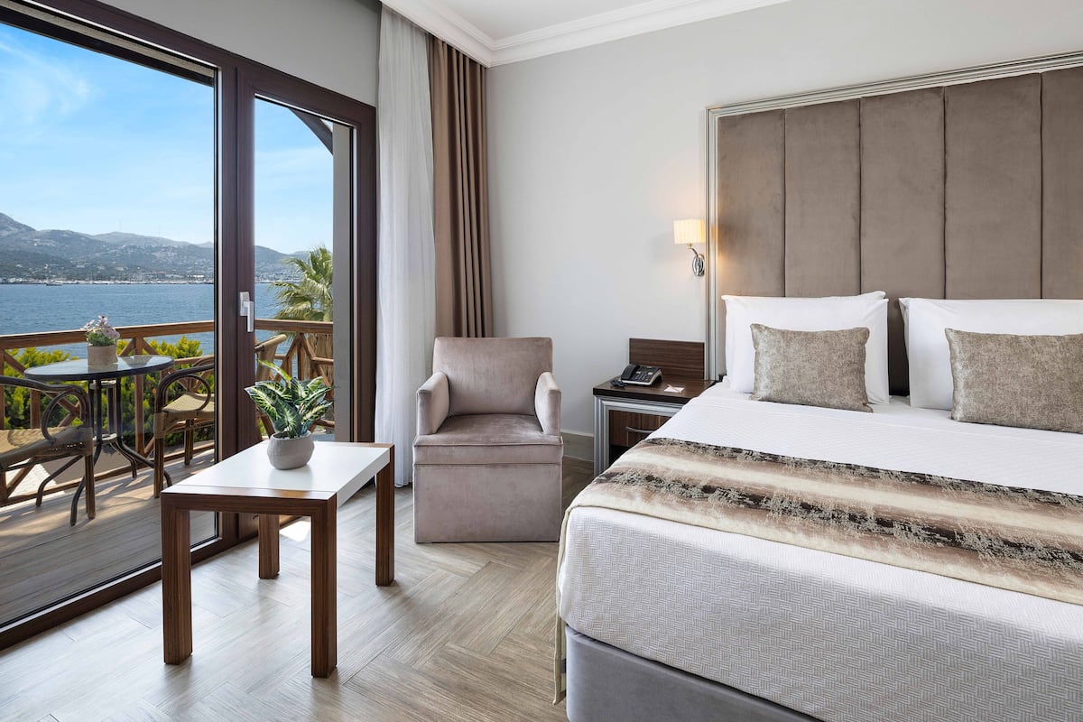 Habitación de hotel con una cama grande, un escritorio, una silla, una mesita con una planta y un balcón con vista al mar y las montañas.