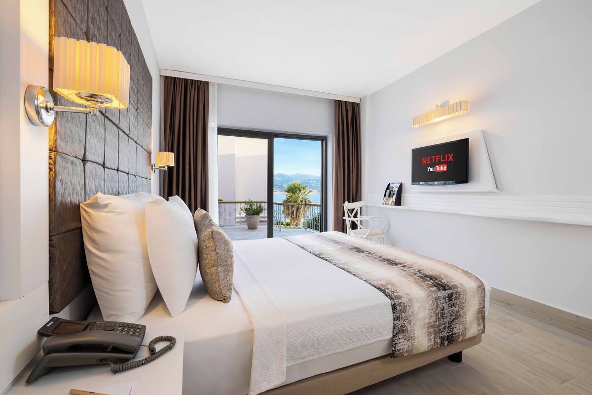 Una habitación de hotel moderna con una cama grande, un teléfono en la mesita de noche, un televisor montado en la pared y vistas al exterior.