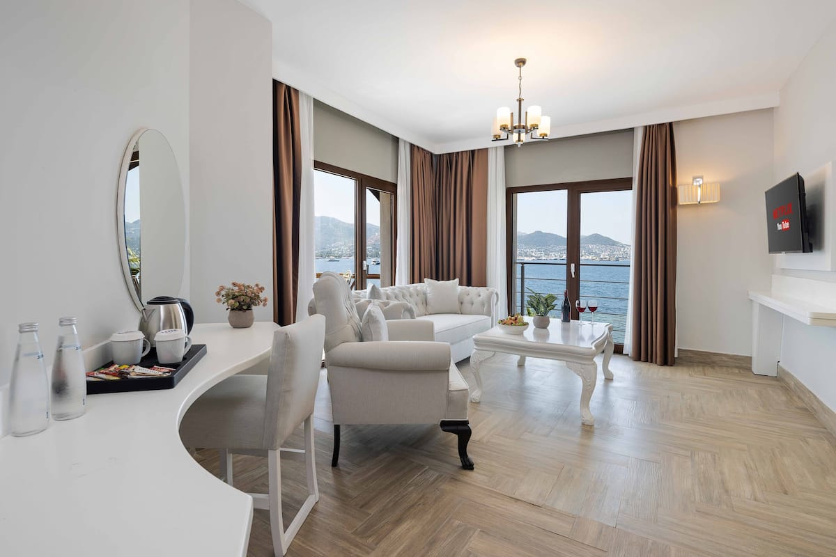 Habitación de hotel moderna con un escritorio blanco curvo, dos sofás blancos, una mesa de centro blanca y un espejo grande. Hay un televisor montado en la pared, una araña y vistas al mar a través de ventanas amplias.