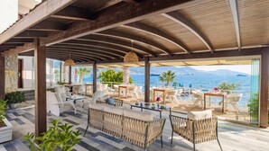 An outdoor lounge area with wicker furniture, a view of the sea, and a wooden ceiling structure.
