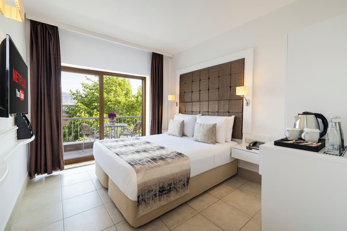 Habitación de hotel con cama, escritorio, cafetera, televisor y balcón con vistas a árboles.