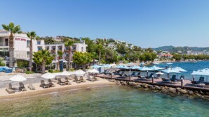 A coastal hotel with a beachside lounge area, clear waters, and a view of the town in the background.