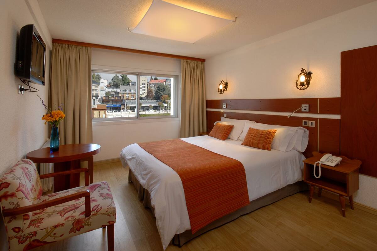 Habitación de hotel con cama, escritorio, silla, televisor, ventana y vistas de la ciudad.