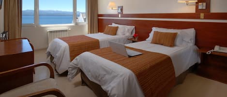 A hotel room with two beds, a desk, a chair, a lamp, and a view of the sea.