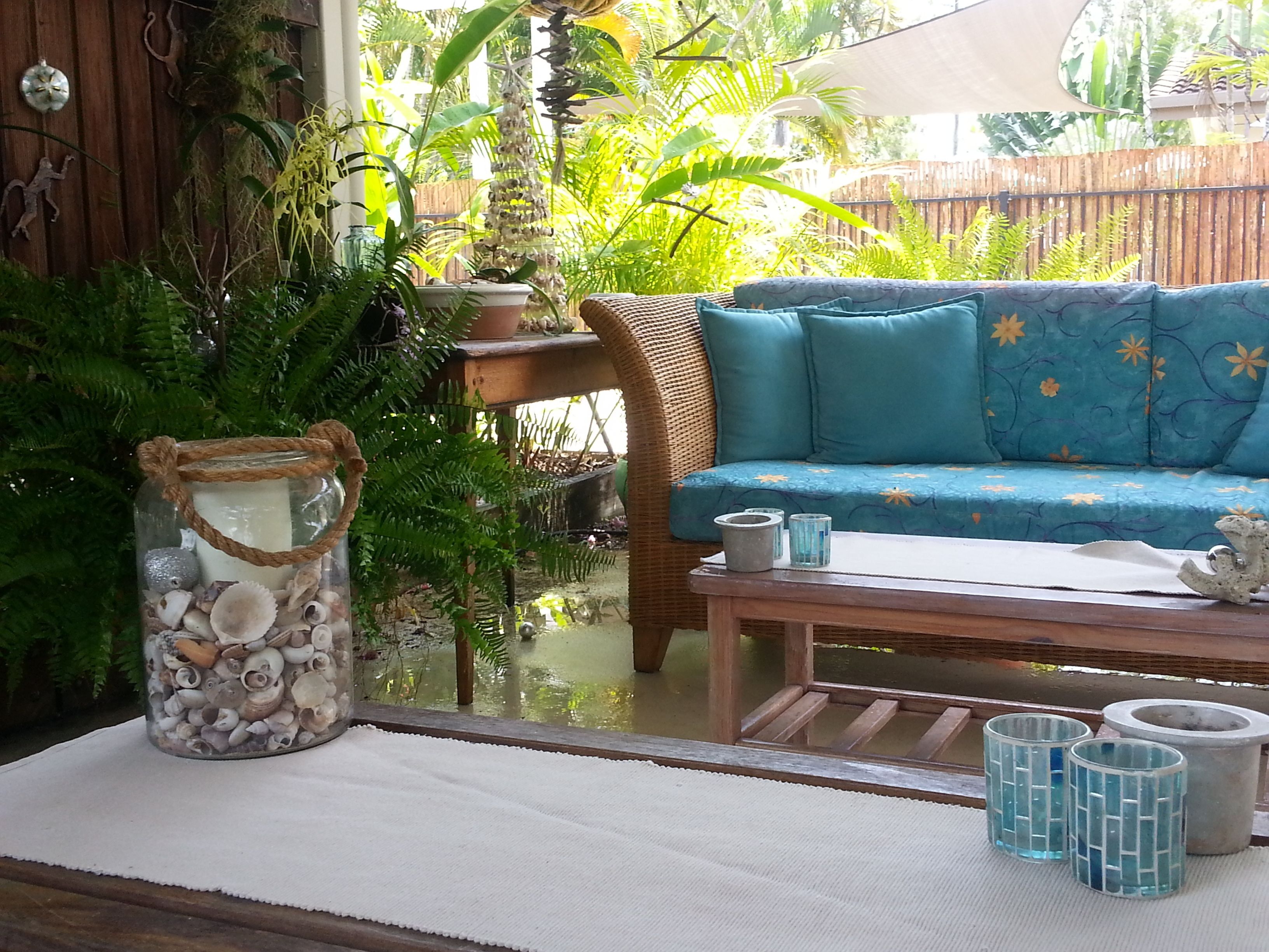 A cozy outdoor seating area with a wicker sofa, a glass vase with seashells, and a small table with two blue glass cups.
