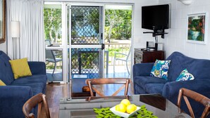 Un salón con dos sofás azules, una mesa de centro de vidrio con limones, una silla de madera, un televisor y un ventanal corredero que da a un espacio al aire libre.