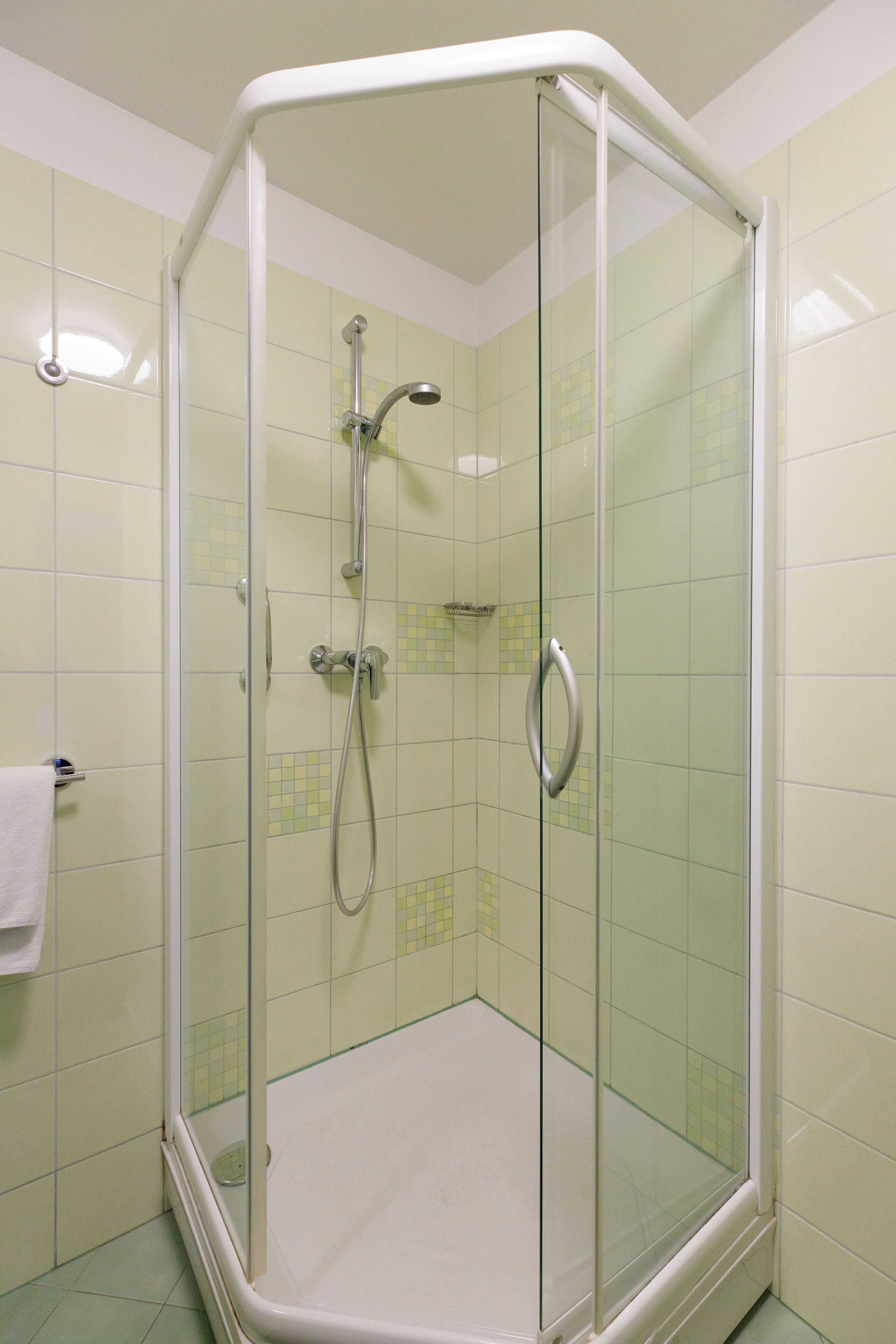 Ducha con mampara de vidrio, puerta blanca con manija, paredes con azulejos y piso blanco.