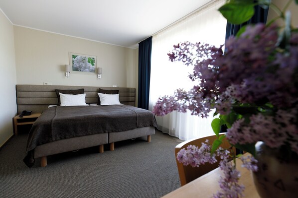 A hotel room with a bed, bedside tables, a nightstand, a wall-mounted lamp, and a window with curtains. - Spa Hotel Ezeri (Sigulda Municipality)