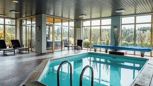 Una piscina interna con pareti in vetro trasparente, lettini prendisole e vista su una zona boscosa.