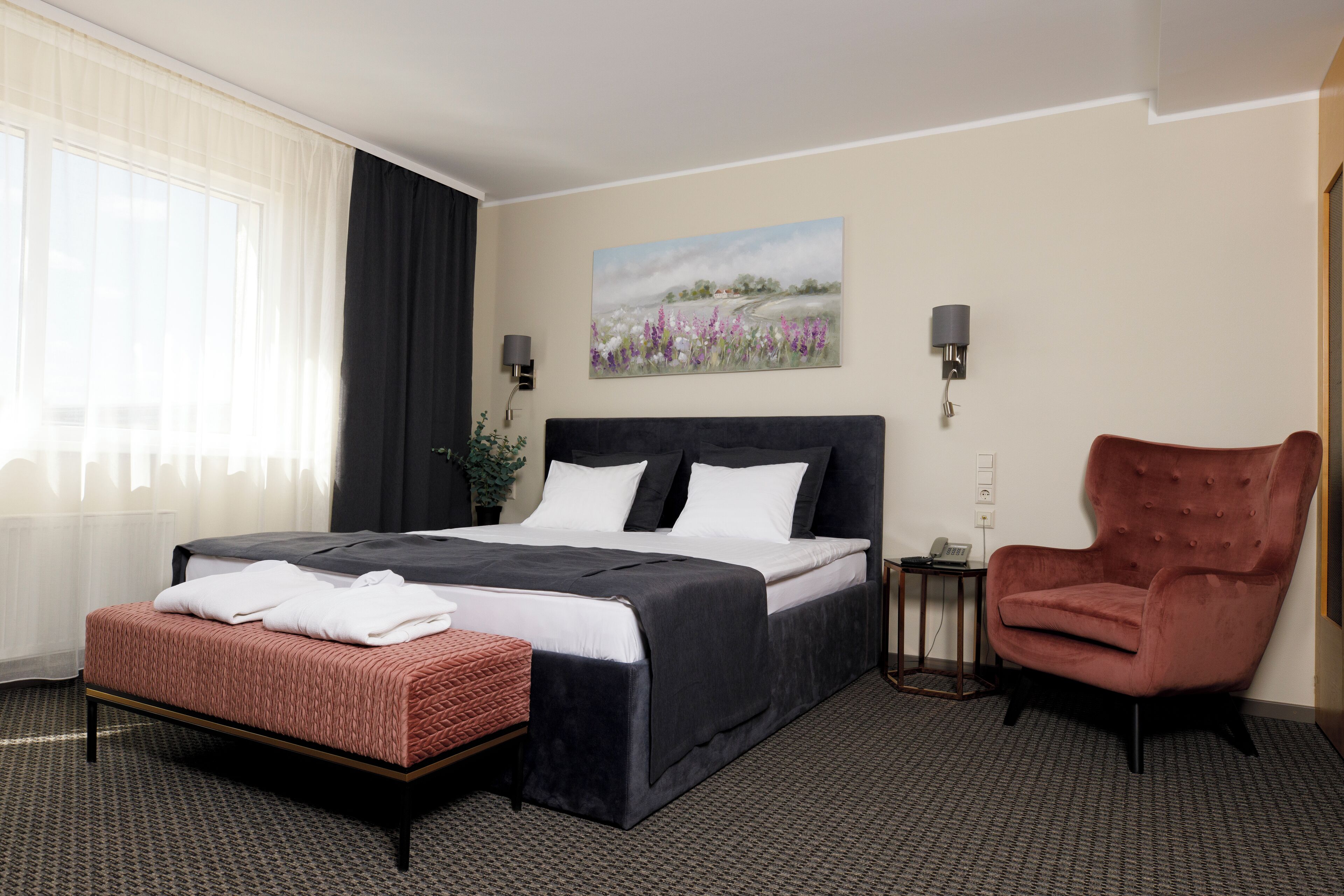 Habitación de hotel con una cama grande, una mesita de noche, una silla, un cuadro en la pared y una ventana con cortinas.