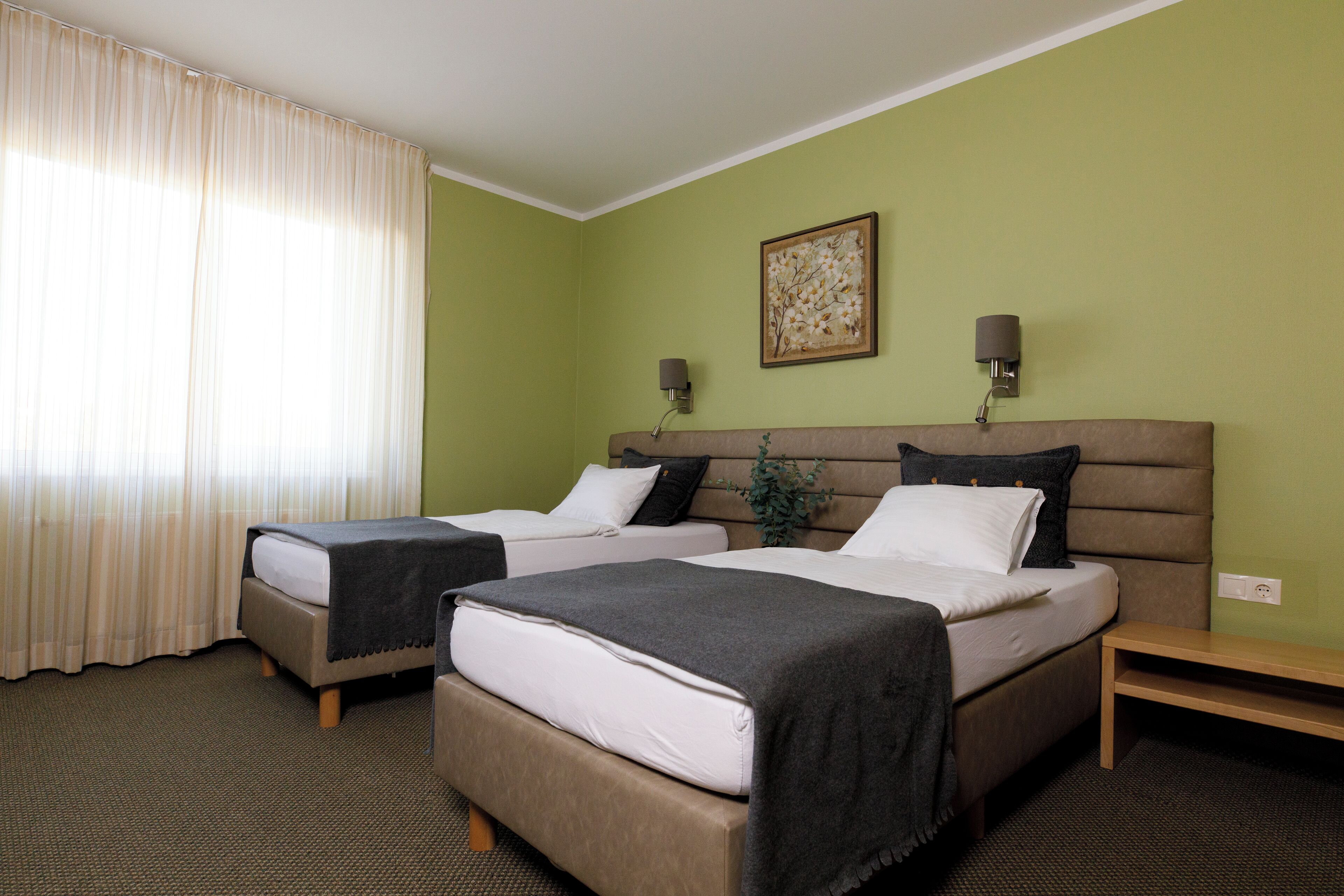 Quarto de hotel com duas camas, uma janela grande com cortinas, uma luminária na parede e uma pequena planta.