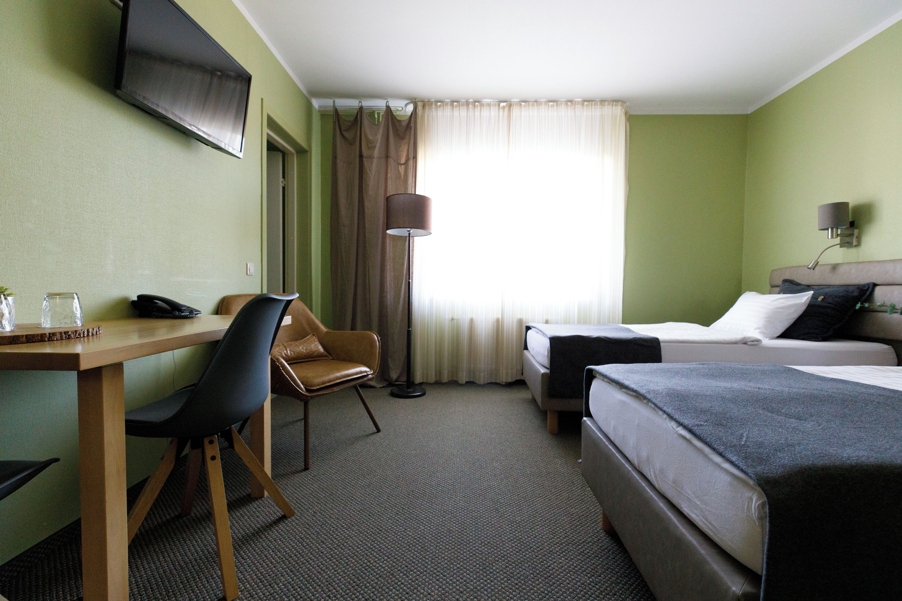 A hotel room with a desk, two beds, a chair, a TV, and a window with curtains.