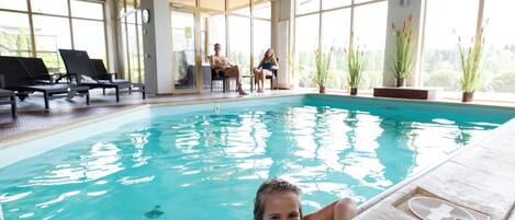 An indoor swimming pool with clear blue water, lounge chairs, and potted plants around the pool area.