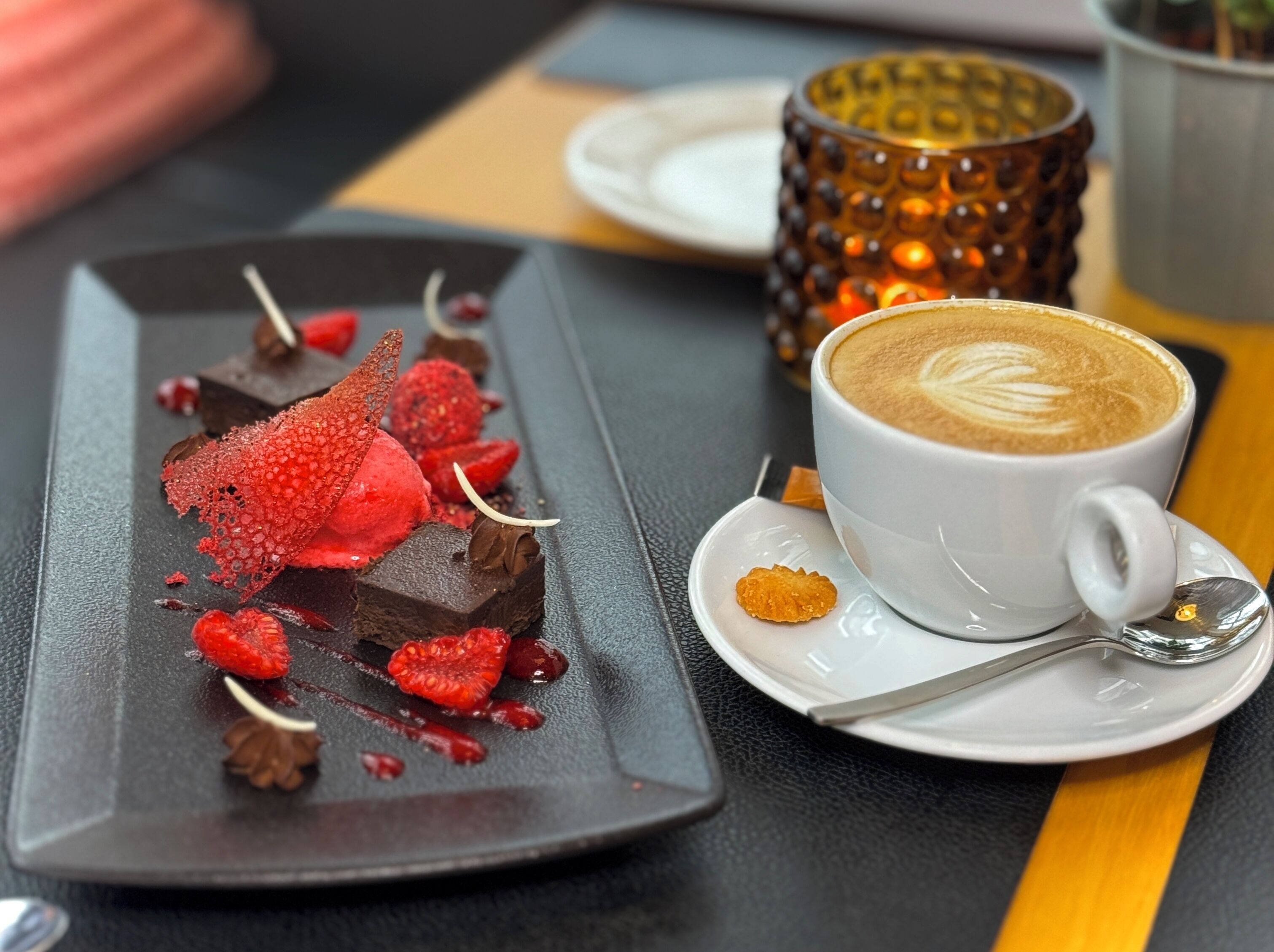 Un plato de postre con fresas y chocolate, una taza de café con arte latte, y una vela decorativa sobre una mesa.