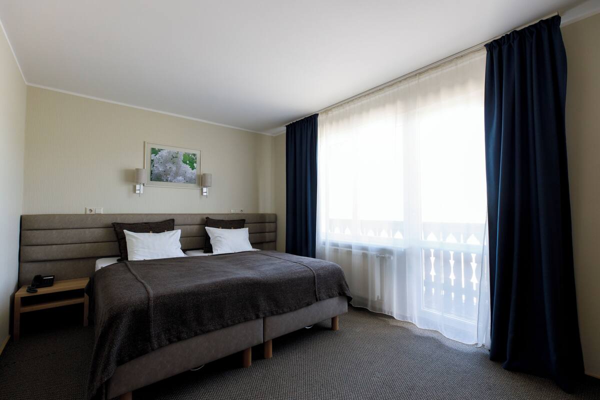 Habitación de hotel con una cama grande, una mesita de noche, una lámpara de pared y una ventana con cortinas.