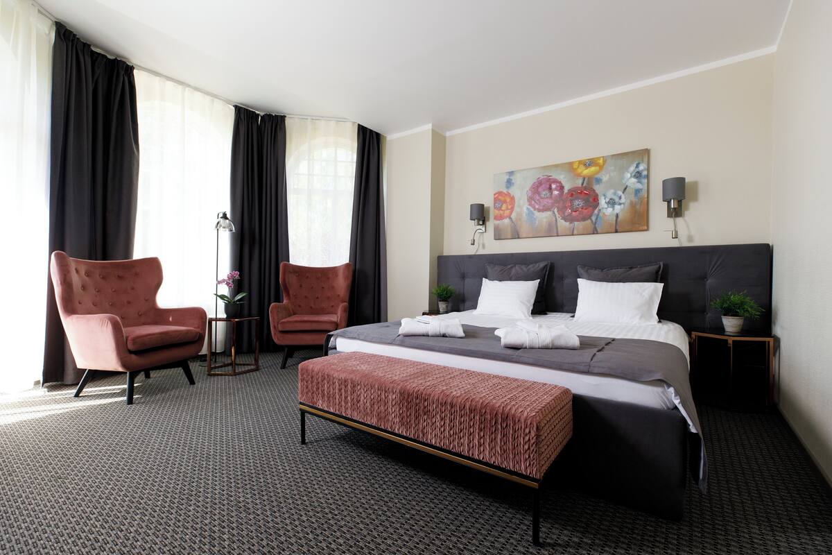 Una habitación de hotel con una cama grande, dos sillones, una mesita con una planta y un cuadro en la pared.