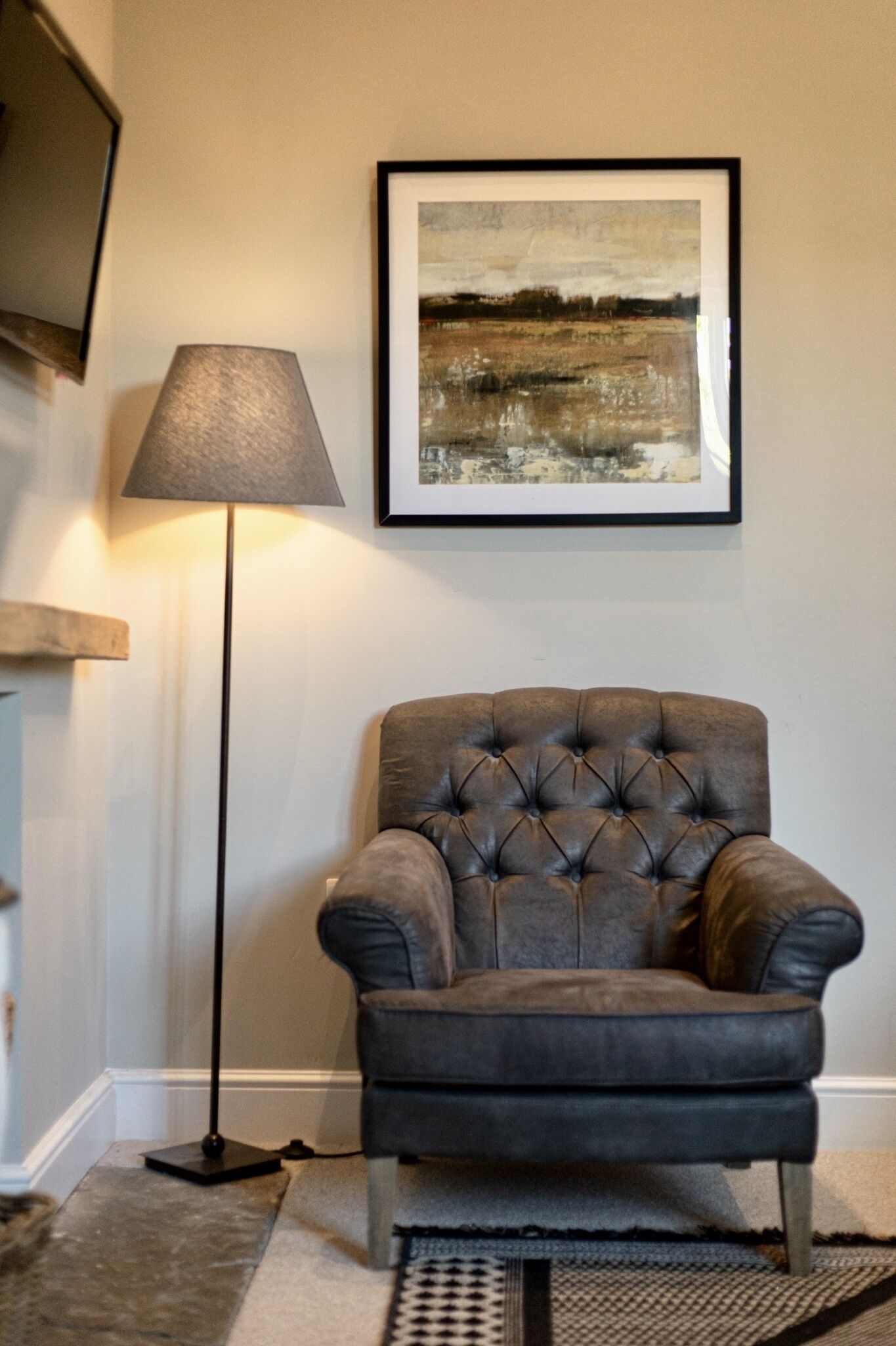 A tufted leather armchair, a floor lamp, and a framed abstract painting on a plain wall.