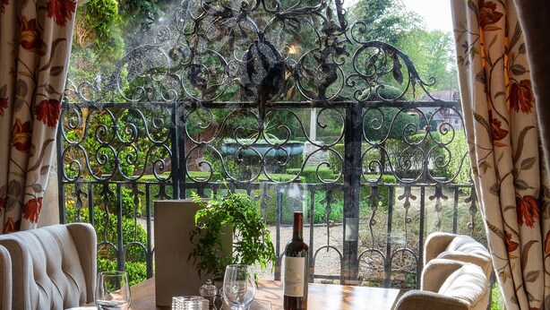 A dining table set with wine glasses, a bottle, and a potted plant, with a view of a garden through a wrought iron gate. - The Five Arrows Hotel (Aylesbury)