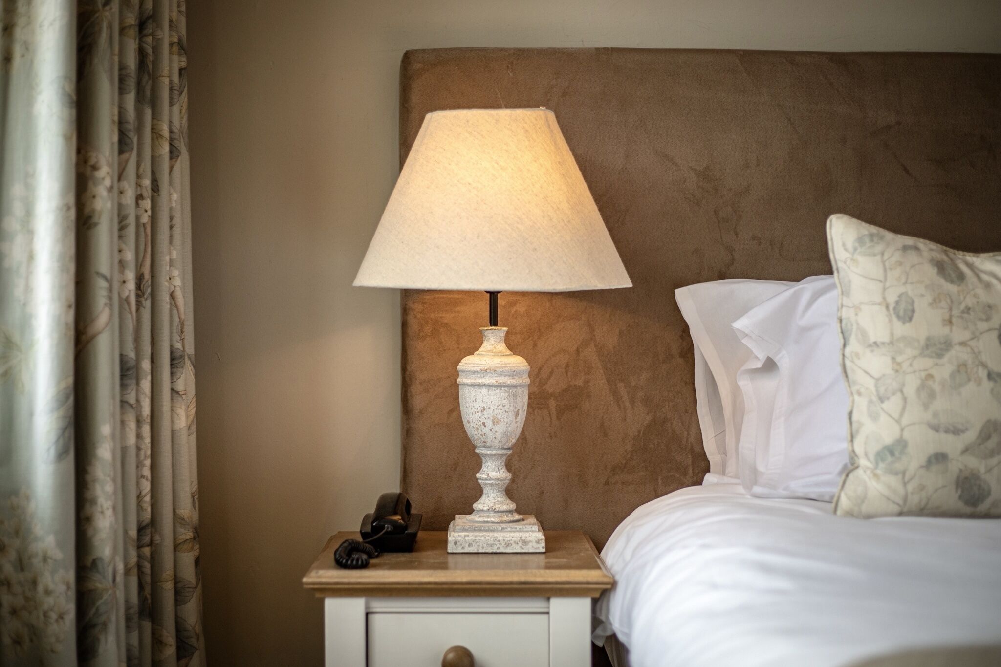 Een net opgemaakt bed met witte lakens, een decoratieve lamp op een nachtkastje en een kussen met bloemenprint.