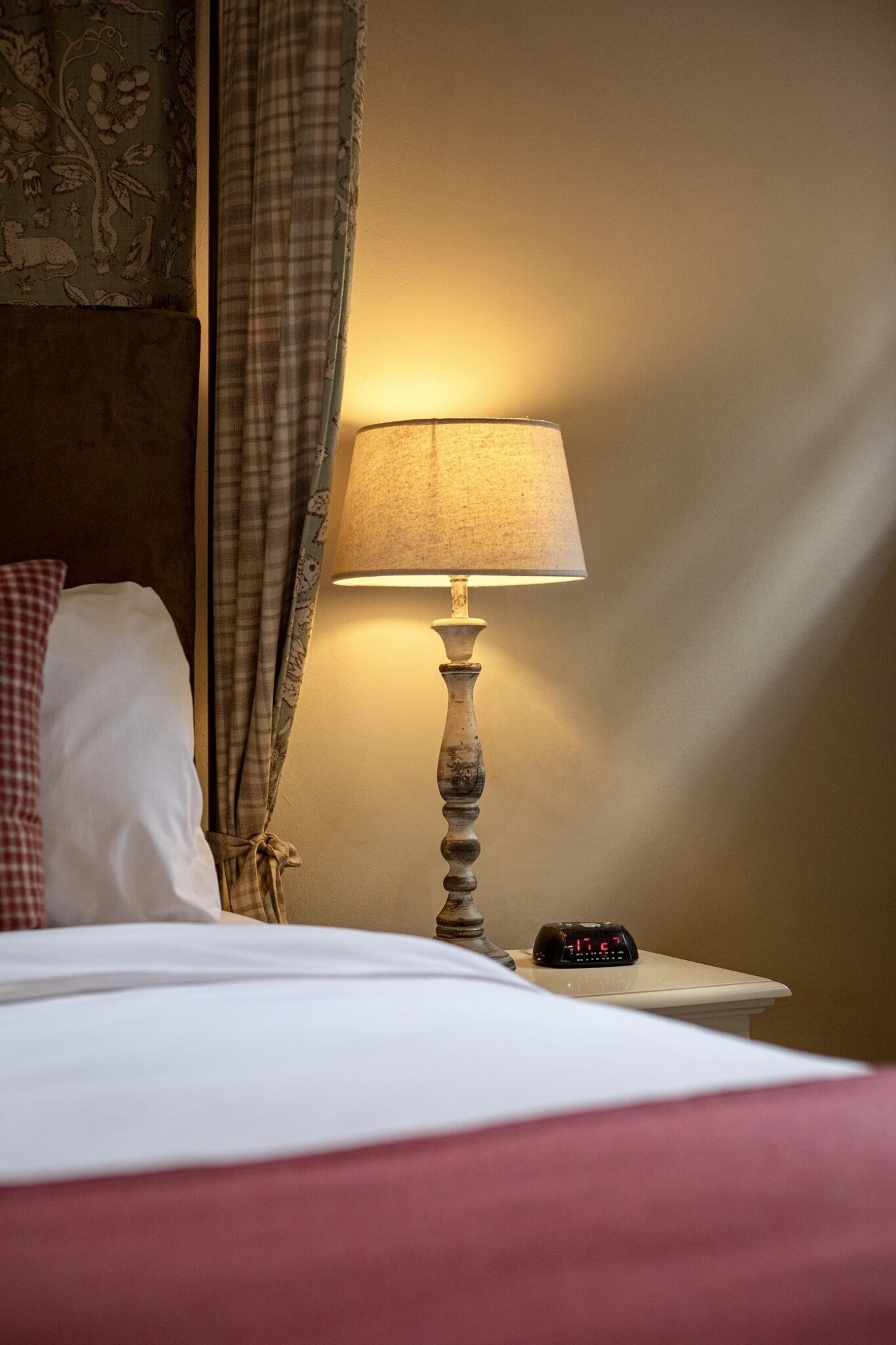 A hotel room with a bed, a bedside lamp, a clock, and a curtain with a floral pattern.