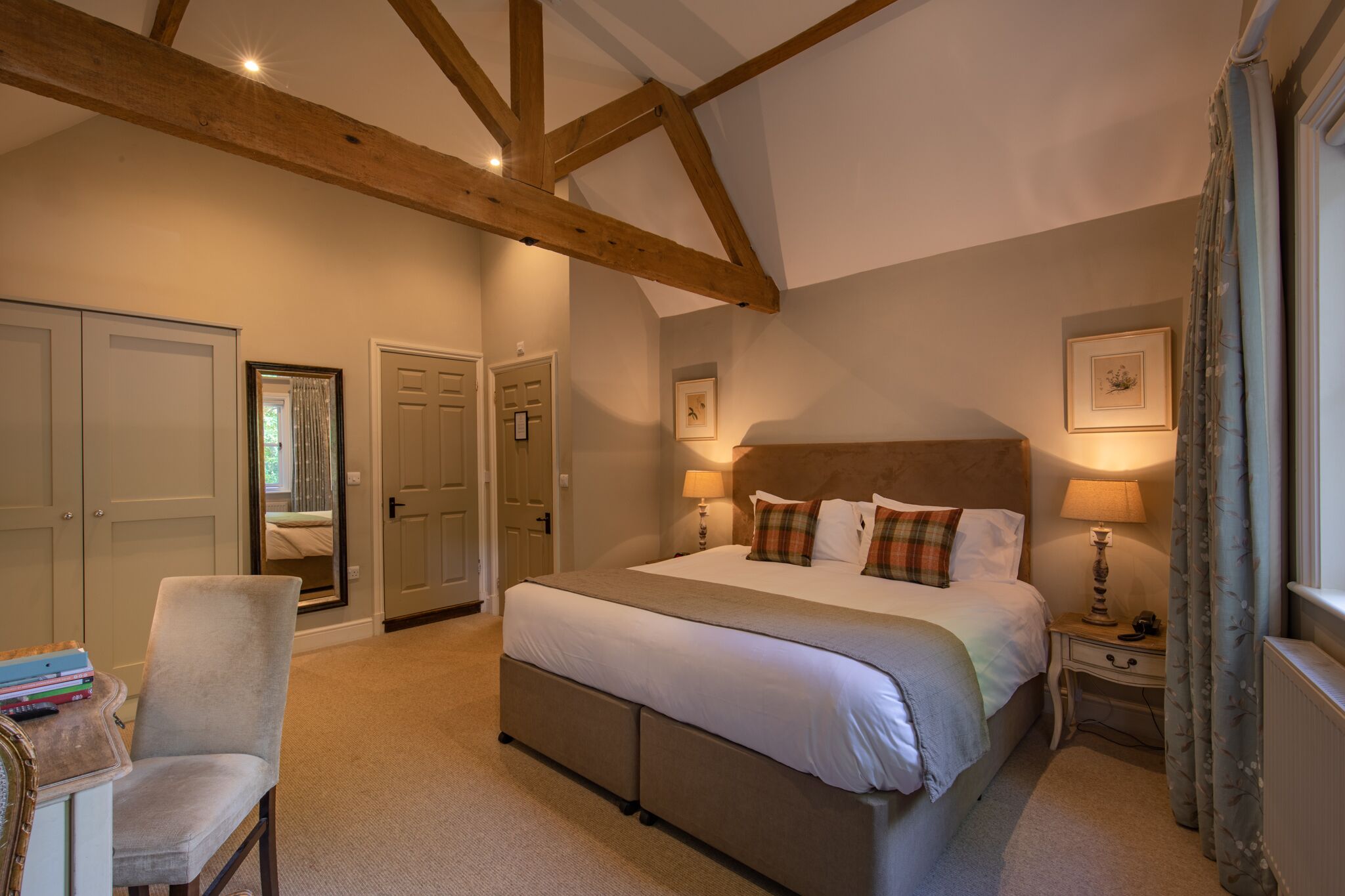 A bedroom with a large bed, wooden beams, a chair, bedside tables, and a window with curtains.