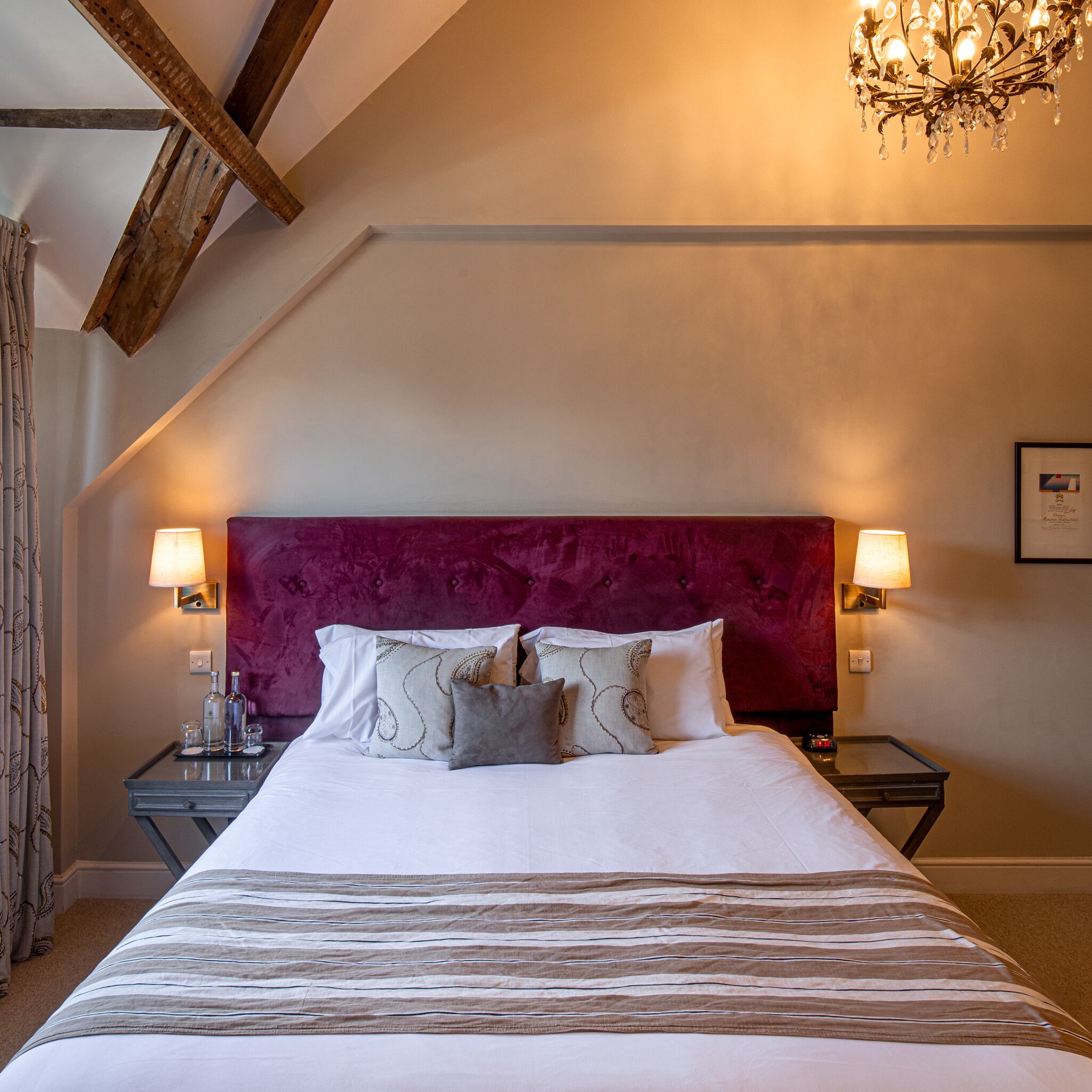 A hotel room with a large bed, a red upholstered headboard, two bedside tables with lamps, and a chandelier.