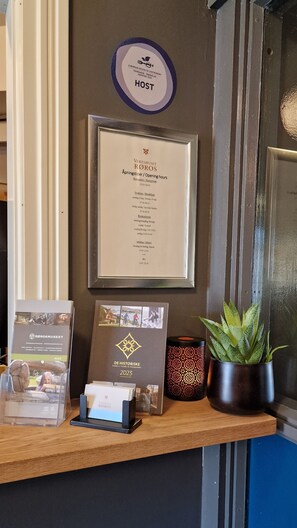A reception desk with a framed menu, a host plaque, a brochure, and a potted plant.