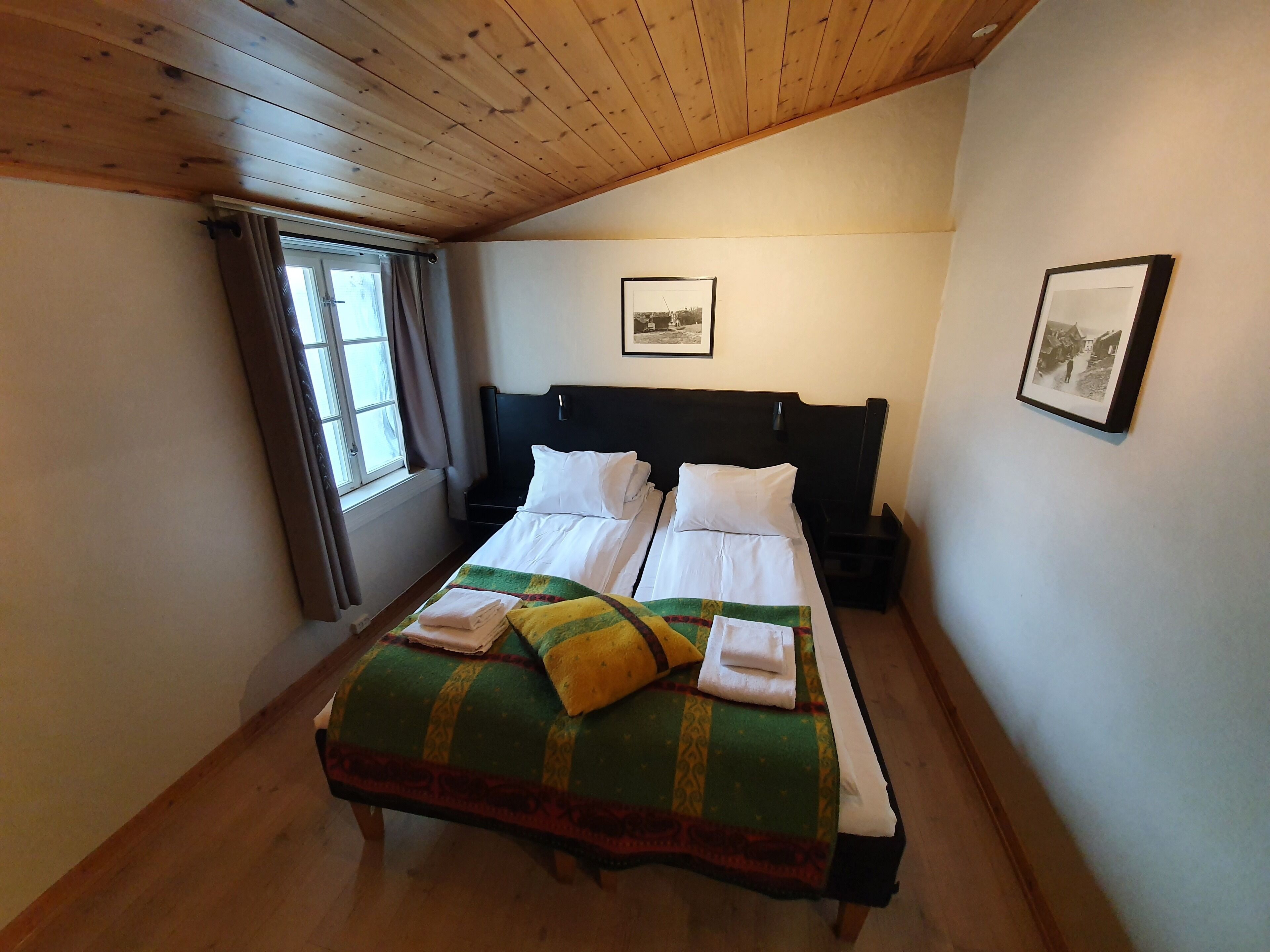 A bedroom with a wooden ceiling, a bed with white linens, a green and yellow blanket, and a window with curtains.