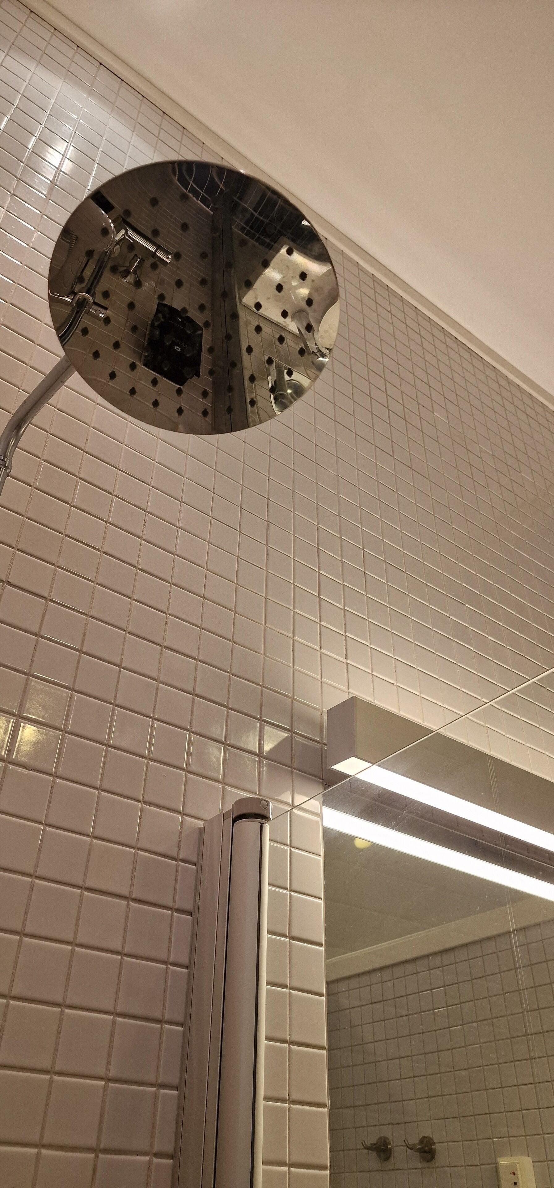 Un baño con paredes de azulejos blancos, un cabezal de ducha y un espejo.