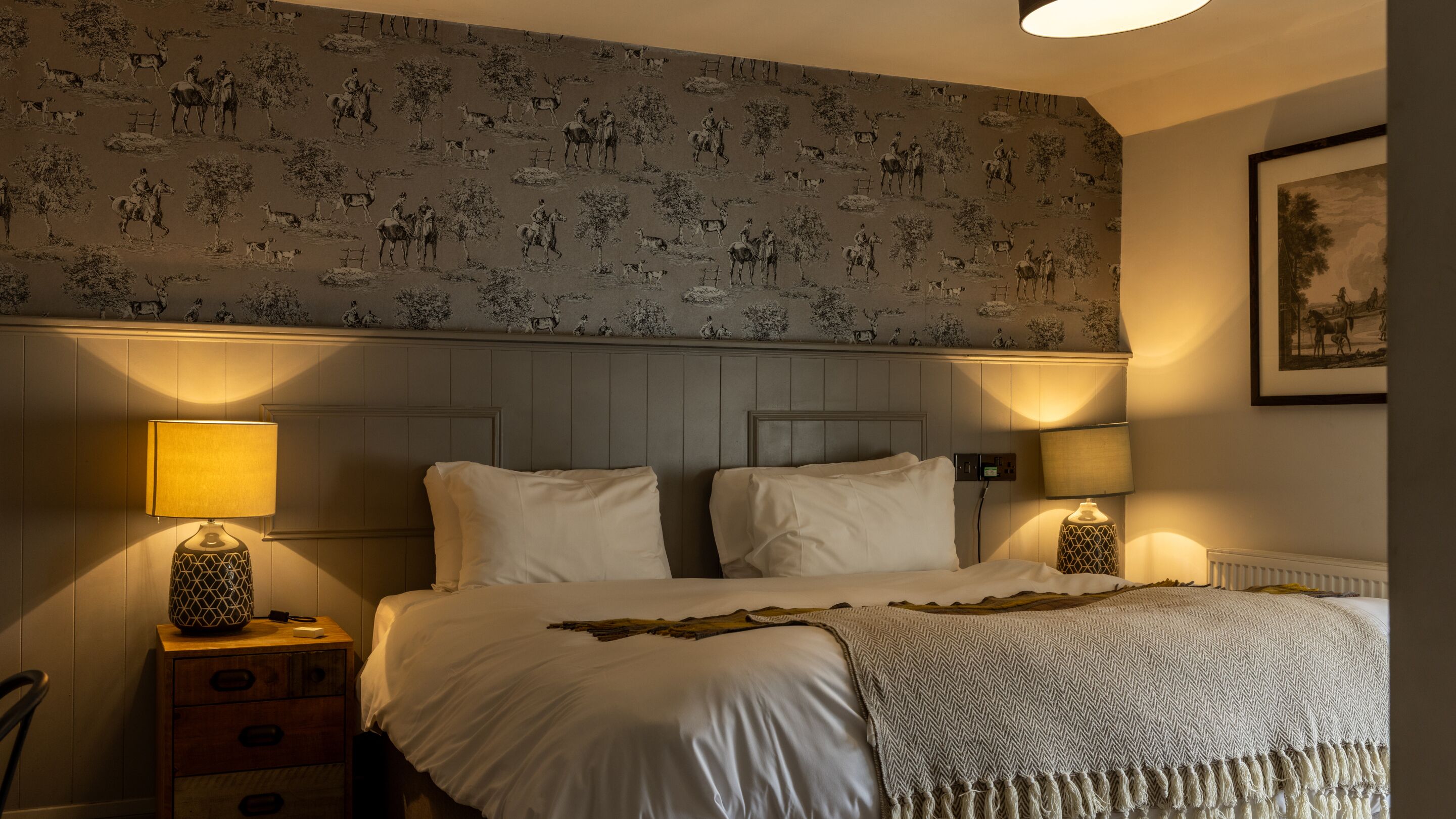 A bedroom with a patterned wallpaper, a wooden headboard, two bedside lamps, a bed with white linens, and a framed picture on the wall.