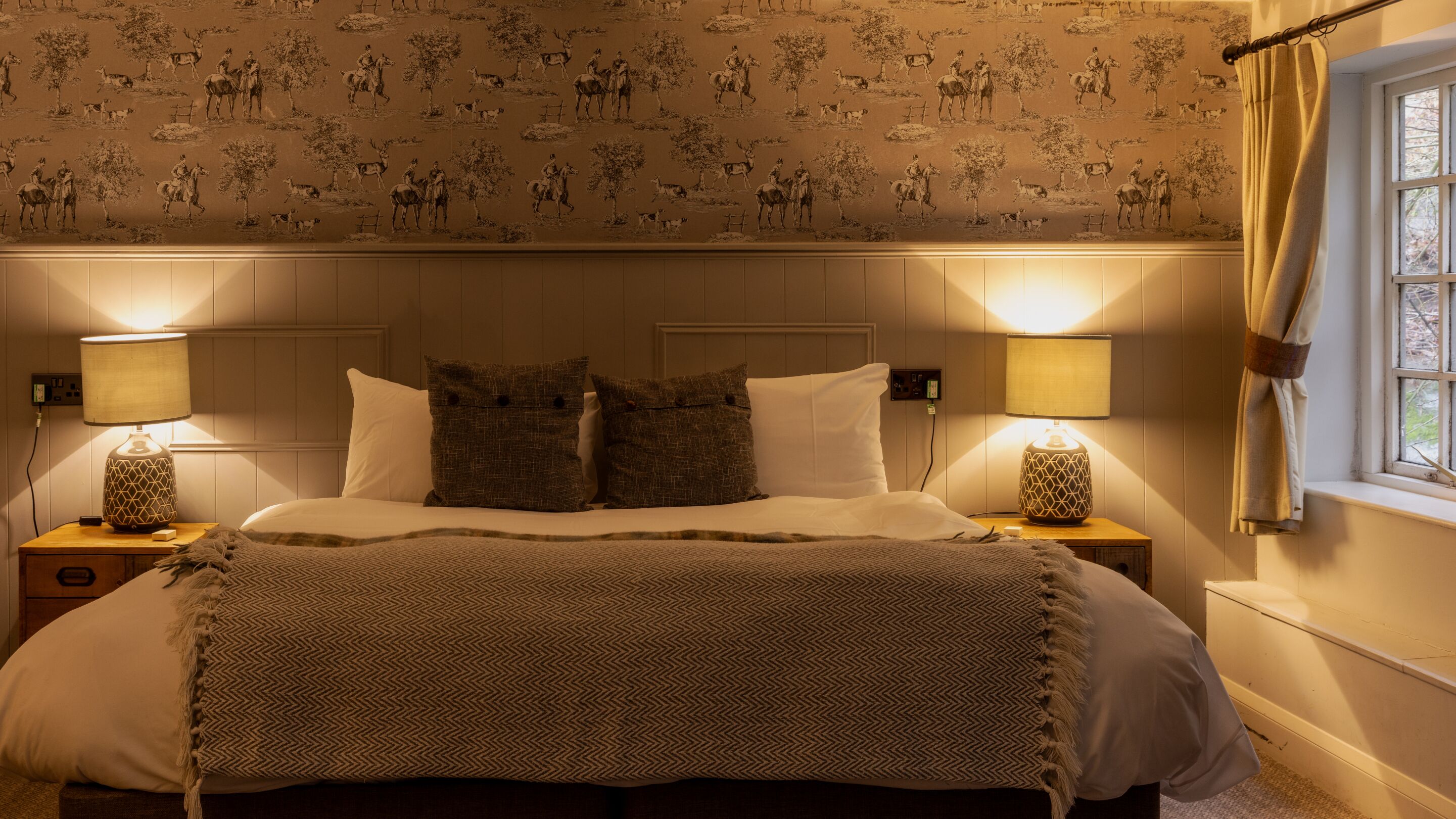 A bedroom with a patterned wallpaper, a bed with a herringbone throw, two bedside lamps, and a window with curtains.